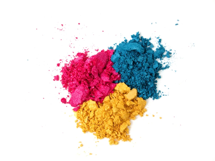Poudres colorées dispersées sur fond blanc: pigments magenta vif, bleu cyan intense et jaune éclatant formant un triangle chromatique