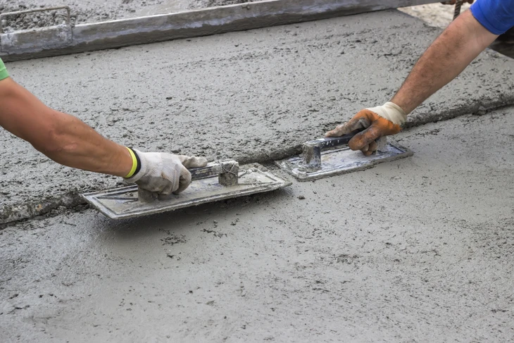 Deux ouvriers du bâtiment travaillent à l'extérieur sur une surface de béton frais, utilisant des truelles de lissage pour égaliser et lisser la surface