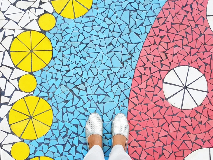 Personne debout sur un sol en mosaïque colorée aux motifs géométriques, portant des chaussures blanches perforées et un pantalon blanc