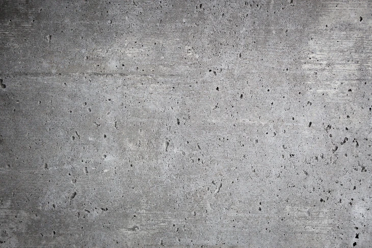 Surface en béton brut gris clair avec texture naturelle présentant de nombreuses micro-fissures, pores et inclusions sombres caractéristiques du matériau coulé