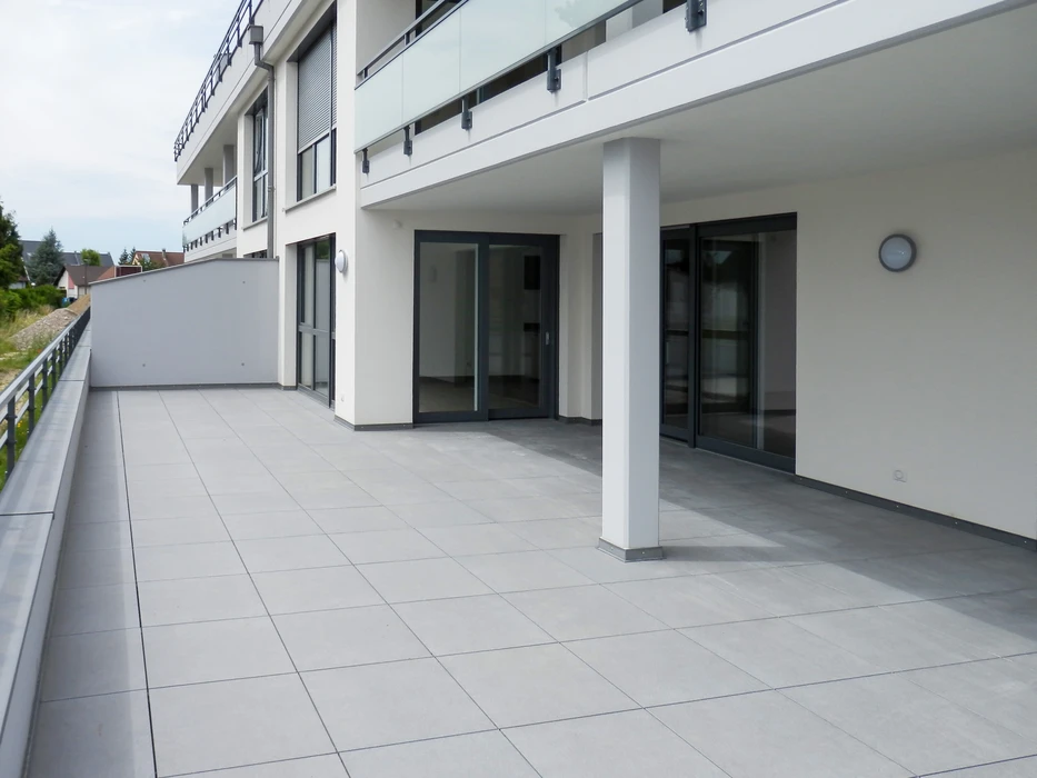 Grande terrasse extérieure moderne en béton, carrelage grès cérame gris clair format 60x60, couverte en partie par un auvent