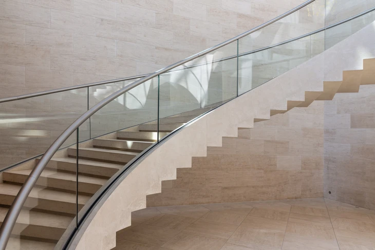 Escalier courbe moderne en pierre naturelle beige avec garde-corps en verre et main courante métallique dans un espace intérieur contemporain aux murs revêtus de travertin