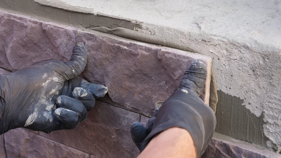 Un homme en chemise à carreaux applique du mastic silicone entre un plan de travail noir et un carrelage mural décoratif dans une cuisine moderne